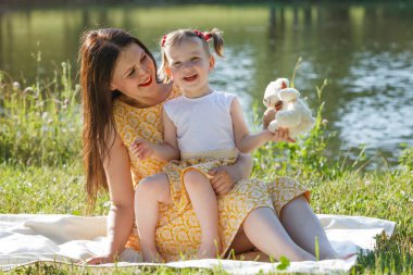 Anne ve kızı üzerinde beyaz bir battaniye oturuyor. Bir beyaz ayı tutan kız. Anne kızı görünüyor. Arka planda göl.