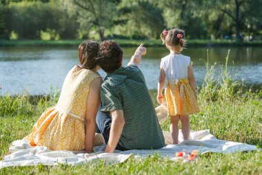 On white plaid sit back parents. Look at the pond. Dad points a finger into the distance. clipart