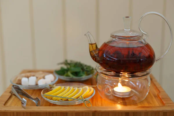 Transparent teapot kettle with tasty black tea on stand with candles. On plates the lemon, sugar, mint and thyme. On wooden background. Shallow depth of field. — Stock Photo, Image