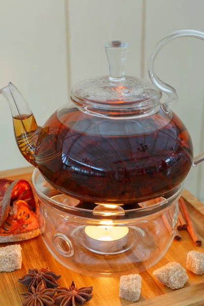 Transparent teapot kettle with tasty black tea on stand with candles. On tray cinnamon, sugar, anise and orange. On wooden background. — Stock Photo, Image