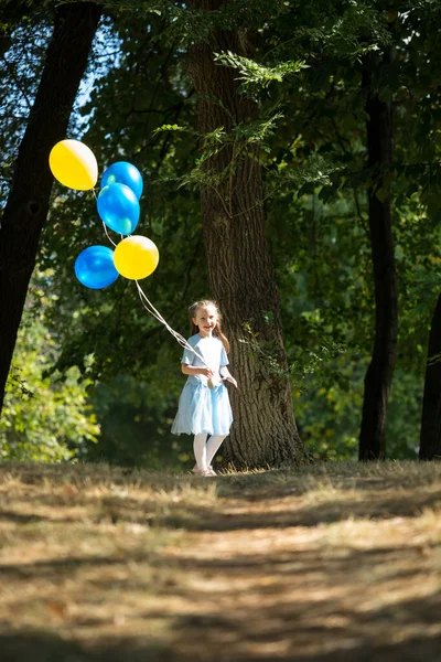 Pequeña chica linda corriendo en el parque con un montón de globos. Se ríe provocativamente. El concepto de una infancia feliz . — Foto de Stock