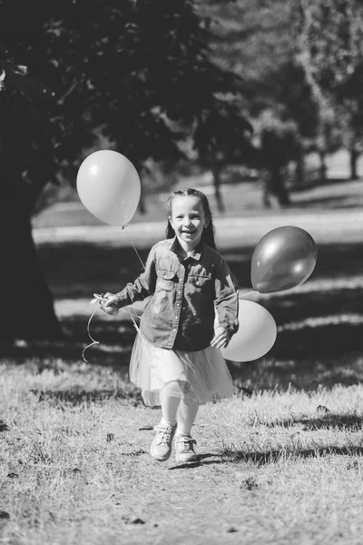 Pequeña chica linda corriendo en el parque con un montón de globos. Se ríe provocativamente. El concepto de una infancia feliz . — Foto de Stock