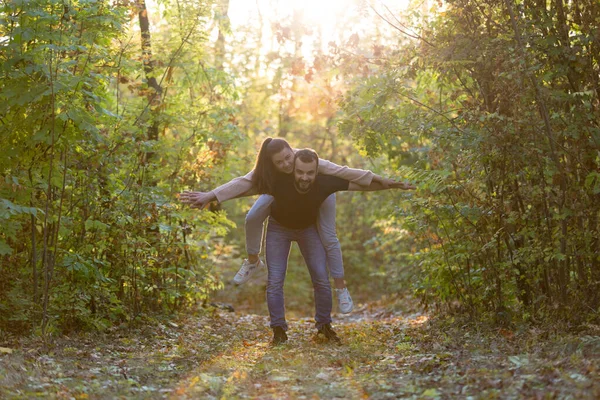 Hermosa pareja joven en el Parque. Un hombre sostiene a una mujer en su espalda, y ellos extienden sus brazos. En el bosque de otoño de fondo. Puesta de sol y resplandor . — Foto de Stock