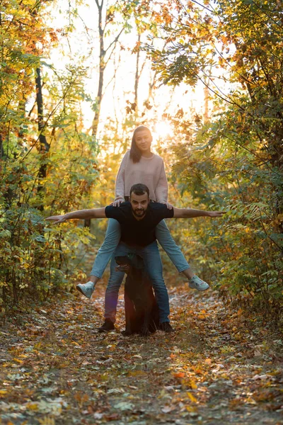 Joven hermosa pareja y su labrador en el Parque. Un hombre sostiene a una mujer en su espalda, y ellos extienden sus brazos. Rayos de sol poniente rompen las ramas de los árboles . — Foto de Stock