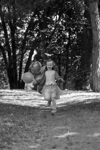 Pequeña chica linda corriendo en el parque con un montón de globos. Se ríe provocativamente. El concepto de una infancia feliz . — Foto de Stock
