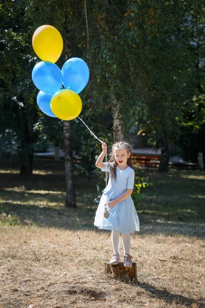 Niña linda en el parque con un montón de globos. Está de pie sobre un muñón. El concepto de una infancia feliz . — Foto de Stock