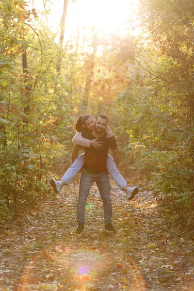 Молода красива пара та їхній лабрадор у парку. Чоловік тримає жінку на спині, і вона обіймає його. Промені встановлення сонця прориваються крізь гілки дерев . — стокове фото