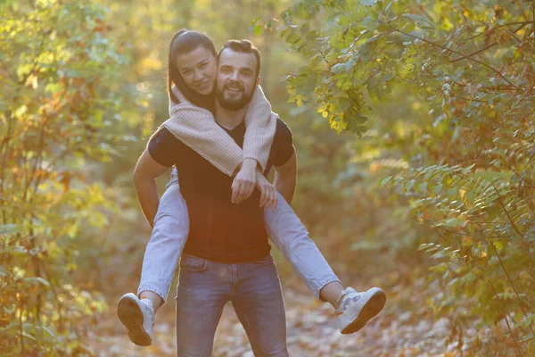 Jeune beau couple et leur labrador dans le parc. Un homme tient une femme sur le dos et elle le serre dans ses bras. Soirée d'automne chaude dans la forêt . — Photo