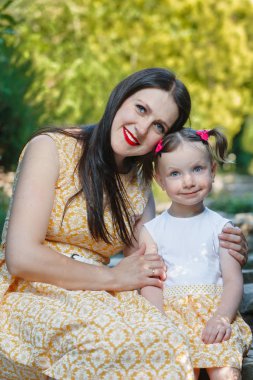 Anne ve kızı parktalar. Bir kadın bir kıza sarılır. Arkaplanda doğa.