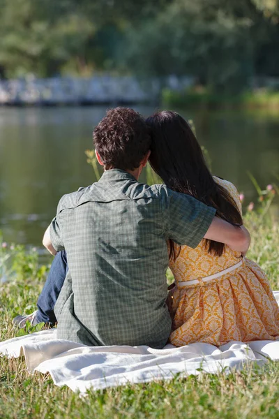 Sobre cuadros blancos se sientan detrás joven pareja. Miran el lago. Un hombre abraza a una mujer . — Foto de Stock