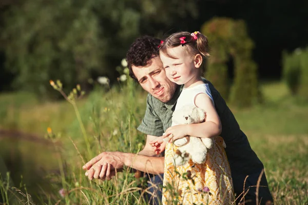 Baba ve kızı göle bakıyor. Kızın elinde oyuncak ayı var. Parlak güneşte gözlerini kısarlar. — Stok fotoğraf