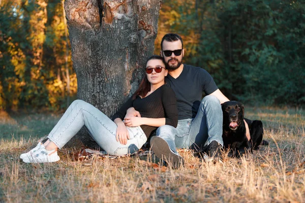 Joven hermosa pareja de su Labrador están sentados en la hierba en el Parque. Cálida tarde de otoño en el bosque. El concepto de una vida familiar feliz en el amor . — Foto de Stock