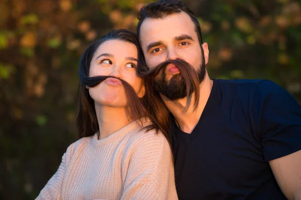 Jong mooi koppel in het park. Ze rotzooien en hebben plezier. Ze maken valse snorren van haar. Ze worden verlicht door een zonnestraal. Op de achtergrond herfstbos. — Stockfoto