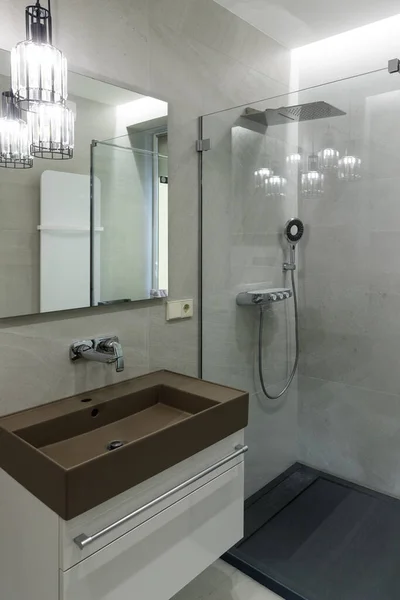 Havalı kahverengi lavabo, krom musluk, büyük ayna. Şeffaf cam duvarlı duş kabini, yağmur duşu ve duş başlığı. Modern banyo tasarımı. Dikey çerçevelenmiş çekim.