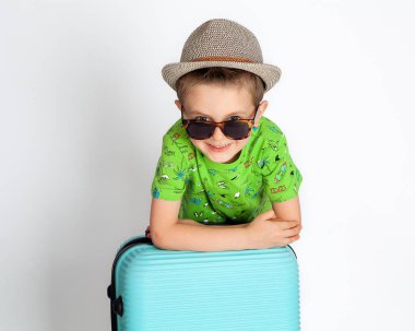 Yolcu çocuk bir bavula yaslanıyor, gözlüklerin üzerinden bakıyor ve gülümsüyor.