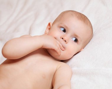 Bebek beyaz yatak örtüsünün üzerinde sırt üstü yatar ve başka yere bakar. Çocukların gözlerini aç. Elini emiyor. Annelik bakımı. Aile ve çocuklar. Ebeveynlik ve annelik.