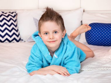Güzel mavi gözlü Avrupalı bir çocuk mavi bir sabahlıkla açık bir yatakta yatıyor. Yüzdükten sonra dinlenin. Hijyen ve çocuk bakımı. Banyo yaptıktan sonra.