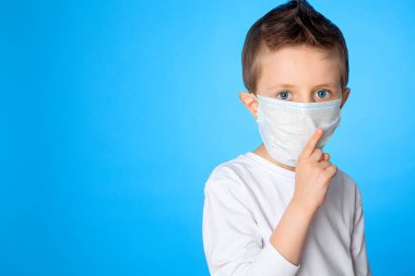 Mavi arka planda tıbbi maske takan beş yaşındaki yakışıklı mavi gözlü bir çocuk parmağını ağzına koydu. Endişe, heyecan, hastalığa yakalanma tehlikesi. Salgınlara karşı mücadele.