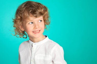 Güzel kıvırcık saçlı, mavi gözlü bir çocuk sinsi sinsi görünüyor ve gülümsüyor. Deniz yeşili arka plana karşı beyaz bir gömlekle. Çocuk duyguları. Çocuklar melektir..