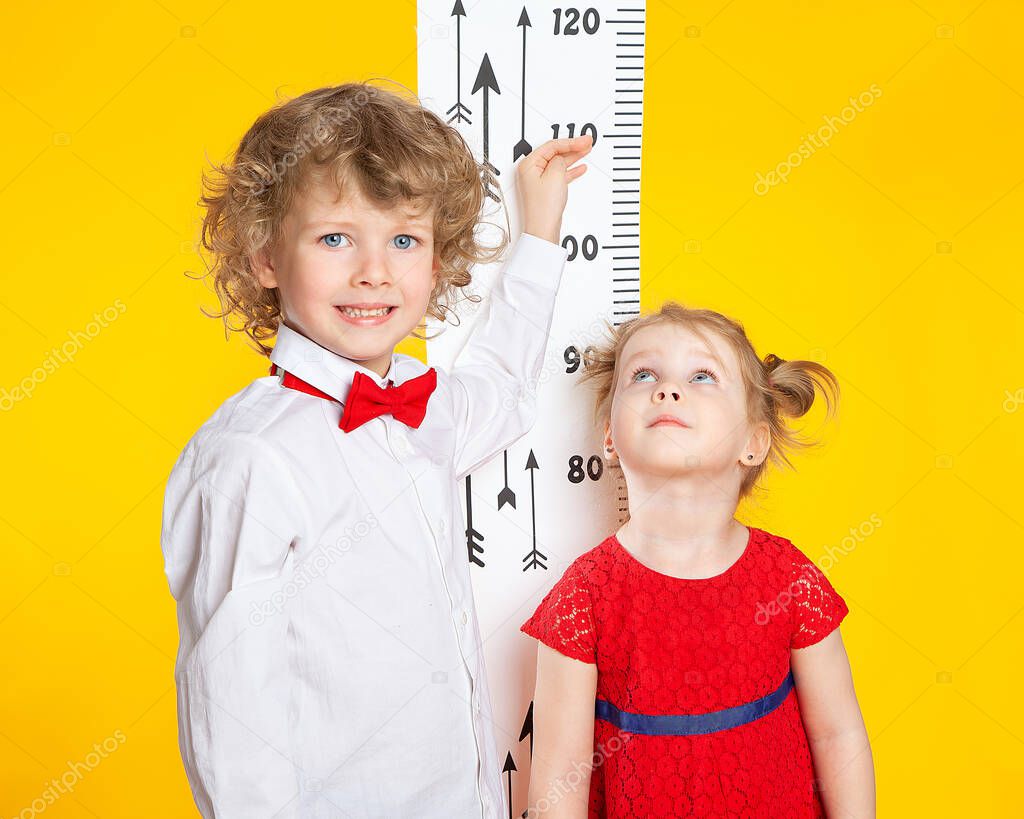 The older brother measures the height of the younger sister using a height meter. Smart children on a yellow background. A boy in a red bow tie. A girl in a red dress. European children.