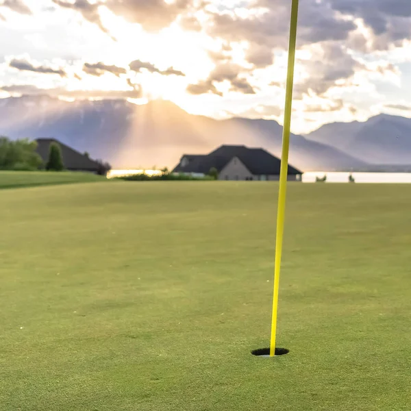 Quadratischer Rahmen gelber Fahnenstab und Loch auf einem Golfplatz gegen Seeberg und bewölkten Himmel — Stockfoto