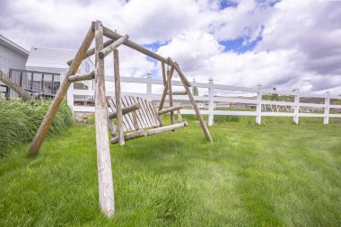 Old wooden garden swing on lush green grasses inside white picket fence clipart
