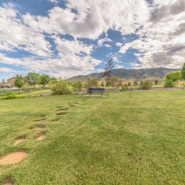 Kare çerçeve, taş basamaklı çimenlik mavi bank, dağ ve bulutlu mavi gökyüzü.