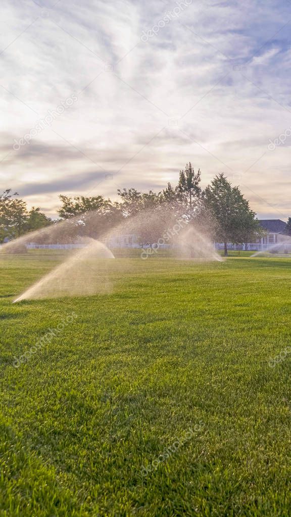 Vertical Sprinklers watering green grassy field with homes and cloudy blue sky background