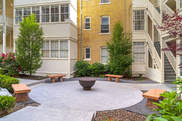 Stone benches around a fire pit outside a residential building on a sunny day. Pathways and plants can also be seen outside the homes with stairways and brick wall.