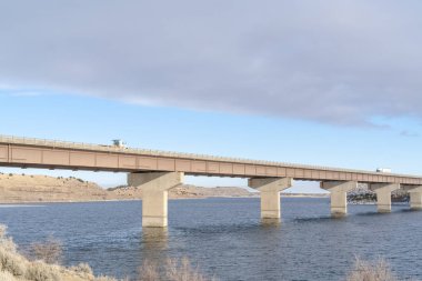 Göl boyunca uzanan köprü, manzaralı tepeler ve kışın bulutlu mavi gökyüzü.