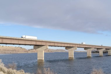 Karlı tepelere ve bulutlu gökyüzüne karşı devasa bir köprüde seyahat eden dev kamyonlar.