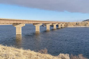 Mavi göl boyunca uzanan köprü karla kaplı tepeler ve bulutlu mavi gökyüzü