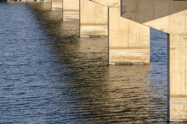 Güneşli göl suyuna karşı bir kiriş köprüsünün altındakileri görür