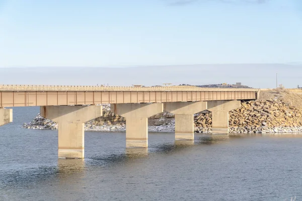 Lago calmo brilhante com longa ponte que leva a um terreno rochoso e nevado — Fotografia de Stock