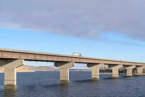 LKW fährt auf Balkenbrücke über See gegen zerklüftetes Land und bewölkten Himmel — Stockfoto