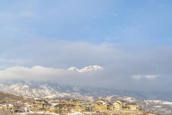 Montagne enneigée partiellement couverte de nuages épais avec des maisons au premier plan — Photo