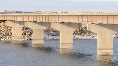 Panorama Parlak sakin göl, uzun köprüyle kayalık ve karlı bir araziye çıkıyor.