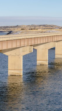 Dikey çerçeve Stringer köprüsü sakin göl üzerinde karlı arazi ve bulutlu gökyüzü manzarasıyla yayılıyor