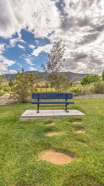 Vertical frame Blue metal bench on vast grassy field with scenic mountain and cloudy sky view