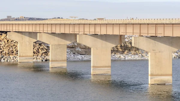 Panorama Shiny calm lake with long bridge that leads to a rocky and snowy terrain — 스톡 사진