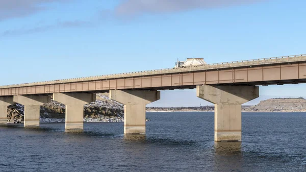 Panorama-LKW fährt auf einer Balkenbrücke über den See gegen zerklüftetes Land und bewölkten Himmel — Stockfoto