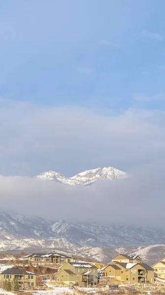 Photo Cadre vertical Montagne enneigée partiellement couverte de nuages épais avec des maisons au premier plan — Photo