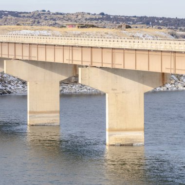 Parlak göl üzerinde çimenli ve karlı manzaralı kare bir köprü.