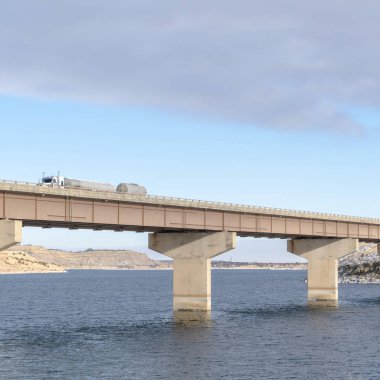 Kare çerçeve Büyük kamyon bulutlu gökyüzünün altında mavi bir gölü geçen bir köprüde seyahat ediyor