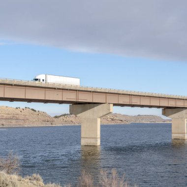 Karlı tepelere ve bulutlu gökyüzüne karşı devasa bir köprüde ilerleyen dev kamyonlar.