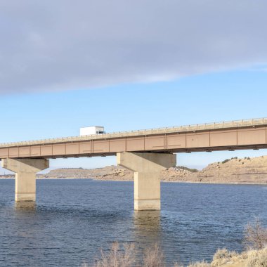Köprüdeki kare kamyon tepelere tepeden bakan mavi göl ve bulutlu gökyüzü.