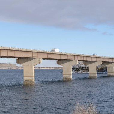 Karlı tepelere ve bulutlu gökyüzüne karşı devasa bir köprüde seyahat eden dev kamyonlar.