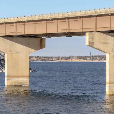 Bulutlu gökyüzüne karşı mavi göl üzerindeki kemerlerle desteklenen bir kiriş köprüsüne odaklan