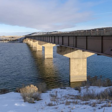 Kış mevsiminde karla kaplı engin sakin bir göl üzerindeki kare köprü.