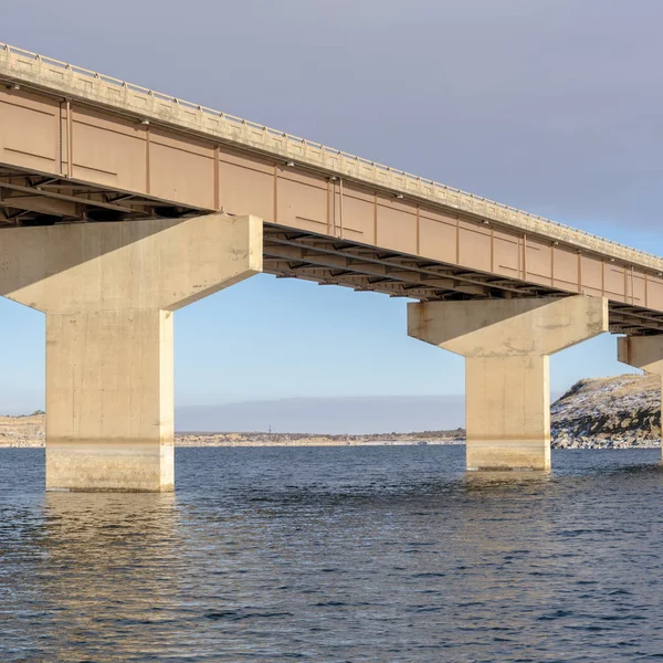 Square frame Stringer bridge spanning over a lake with view of snowy terrain and cloudy sky